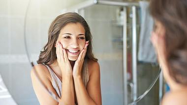 Eine Frau fasst sich lächelnd vor dem Spiegeln ins Gesicht - Foto: istock_Charday Penn