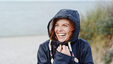 Frierende Frau im Mantel hält lachend die Kapuze fest - Foto: Istock/stockfour