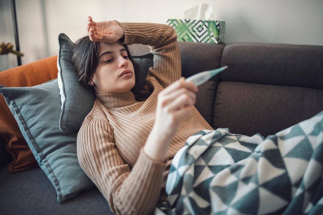 Eine Frau liegt krank auf dem Sofa und misst ihr Fieber