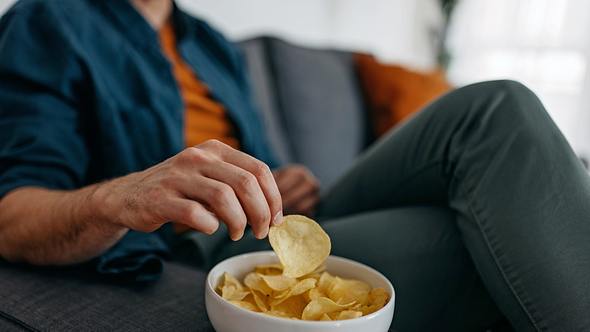 Mann auf Sofa greift in Schale mit Chips - Foto: iStock/supersizer