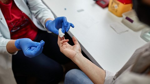 Nahaufnahme von Händen einer Ärztin mit blauen Schutzhandschuhen, die einer Person einen Tupfer an den Finger hält - Foto: iStock/﻿vgajic