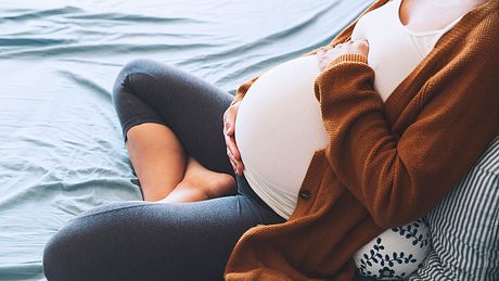 Eine Schwangere sitzt auf dem Sofa und umfasst ihren Bauch - Foto: iStock/NataliaDeriabina