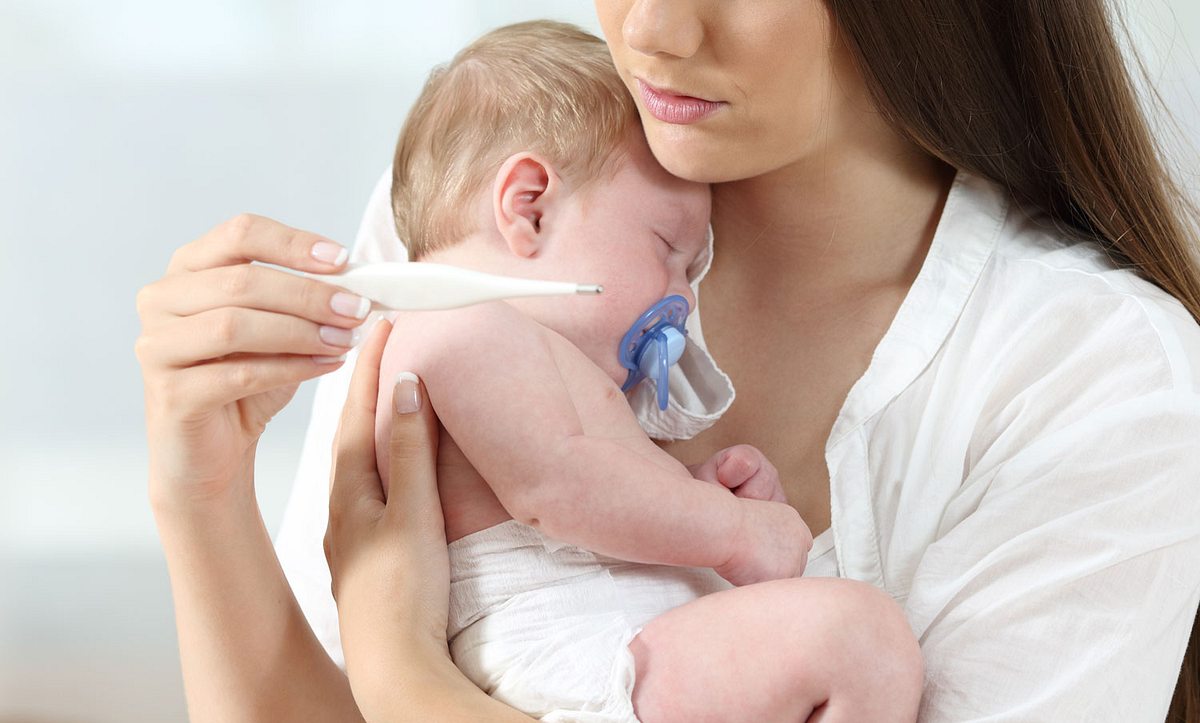 Eine Mutter hat ihr krankes Baby auf dem Arm und schaut auf ein Fieberthermometer