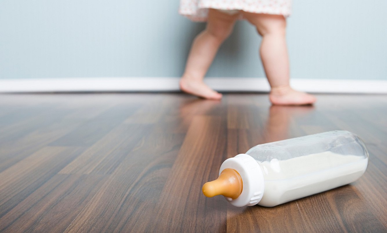 Eine Babyflasche liegt auf dem Boden, dahinter läuft ein Baby