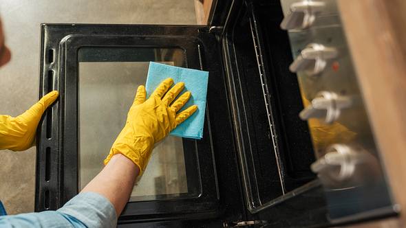 Hände einer Frau mit Handschuhen; sie wischt die Backofentür mit einem Tuch ab - Foto: iStock/Zinkevych