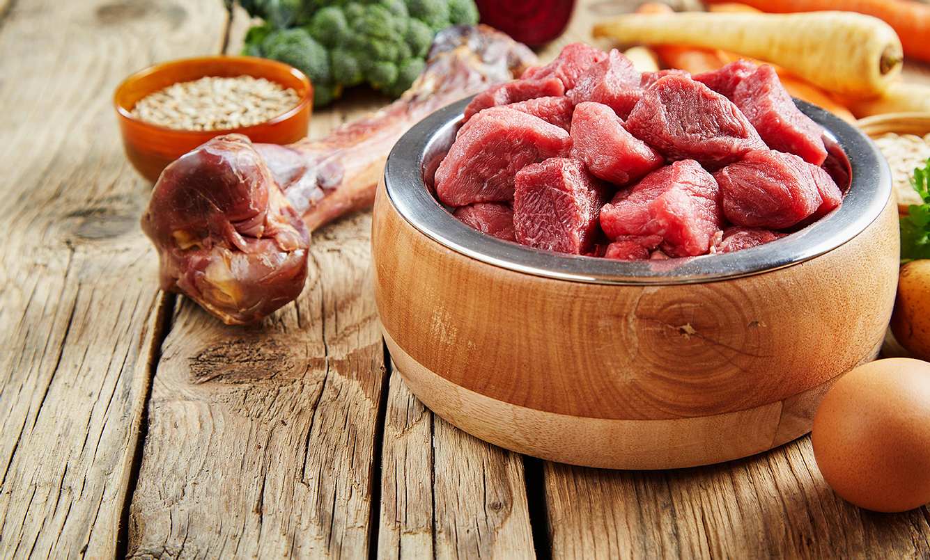 Holzschlüssel mit roh geschnittenem Fleisch steht auf einem Holztisch mit anderen Lebensmitteln im Hintergrund