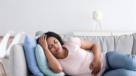 Frau liegt mit Bauchschmerzen auf dem Sofa - Foto: iStock/Prostock-Studio