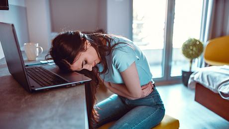 Eine Frau sitzt mit Bauchschmerzen am Schreibtisch - Foto: iStock_AleksandarGeorgiev
