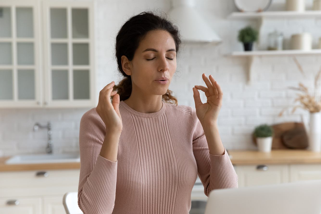 Frau sitzt mit geschlossenen Augen in der Küche, macht Yoga-Finger und atmet bewusst
