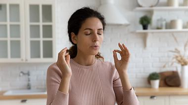 Frau sitzt mit geschlossenen Augen in der Küche, macht Yoga-Finger und atmet bewusst - Foto: iStock/fizkes