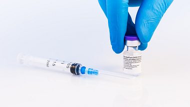 Spritze auf einem weißen Untergrund, daneben hält eine Hand mit Schutzhandschuhen eine Ampulle mit Impfstoff - Foto: istock/carmengabriela