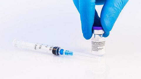 Spritze auf einem weißen Untergrund, daneben hält eine Hand mit Schutzhandschuhen eine Ampulle mit Impfstoff - Foto: istock/carmengabriela