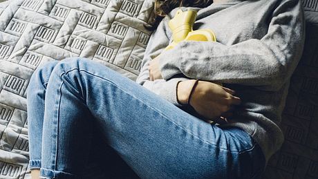 Eine Frau liegt mit einer Wärmflascheauf dem Bett und hält sich den Bauch - Foto: iStock/Carlo107
