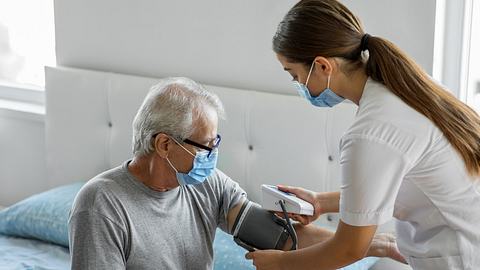 Eine Ärztin prüft den Blutdruck eines Patienten, beide tragen Mundschutz - Foto: iStock-1282000675 bymuratdeniz