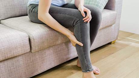 Unterkörper einer Frau in grauer Jeans auf dem Sofa; sie greift sich an den Unterschenkel - Foto: iStock/prpicturesproduction