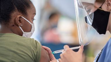 Mädchen mit medizinischer Maske erhält von Ärztin in Schutzkleidung Impfspritze in den Oberarm - Foto: istock/FatCamera