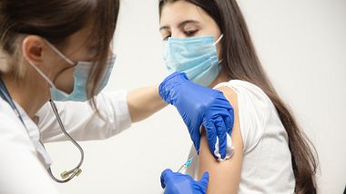 Ärztin mit Schutzmaske und -handschuhen gibt junger Frau mit weißem Shirt und medizinischer Maske eine Infektion - Foto: istock/master1305