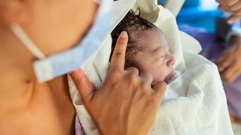 Frau mit Maske hält Säugling im Arm - Foto: iStock / Kemal Yildirim