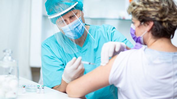 Arzt in Schutzkleidung setzt eine Spritze in den Oberarm einer Patientin mit Maske - Foto: istock/zoranm