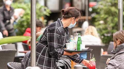 Eine Kellnerin mit Maske bedient einen Gast im Außenbereich eines Restaurants - Foto: IMAGO_Ralph Peter