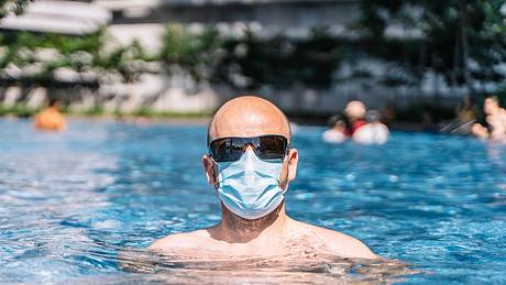 Mann mit Mund-Nasen-Schutz und Sonnenbrille im Pool - Foto: iStock / riderfoot