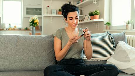 Frau sitzt mit Lanzette auf der Couch - Foto: Istock/PixelsEffect