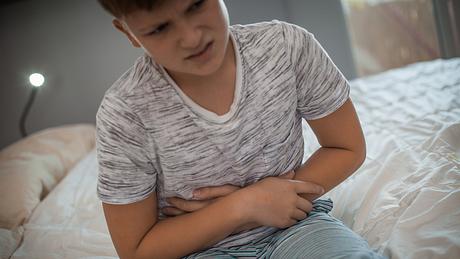 Junge sitzt auf dem Bett und hält sich mit schmerzverzerrtem Gesicht den Bauch - Foto: iStock-1061890064 vitapix