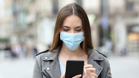 Frau mit Maske guckt auf der Straße auf ihr Handy - Foto: iStock/ AntonioGuillem