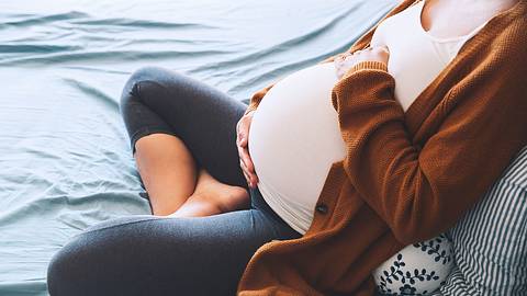 Hochschwangere Frau sitzt im Schneidersitz auf dem Sofa und hält sich den Bauch - Foto: iStock/NataliaDeriabina