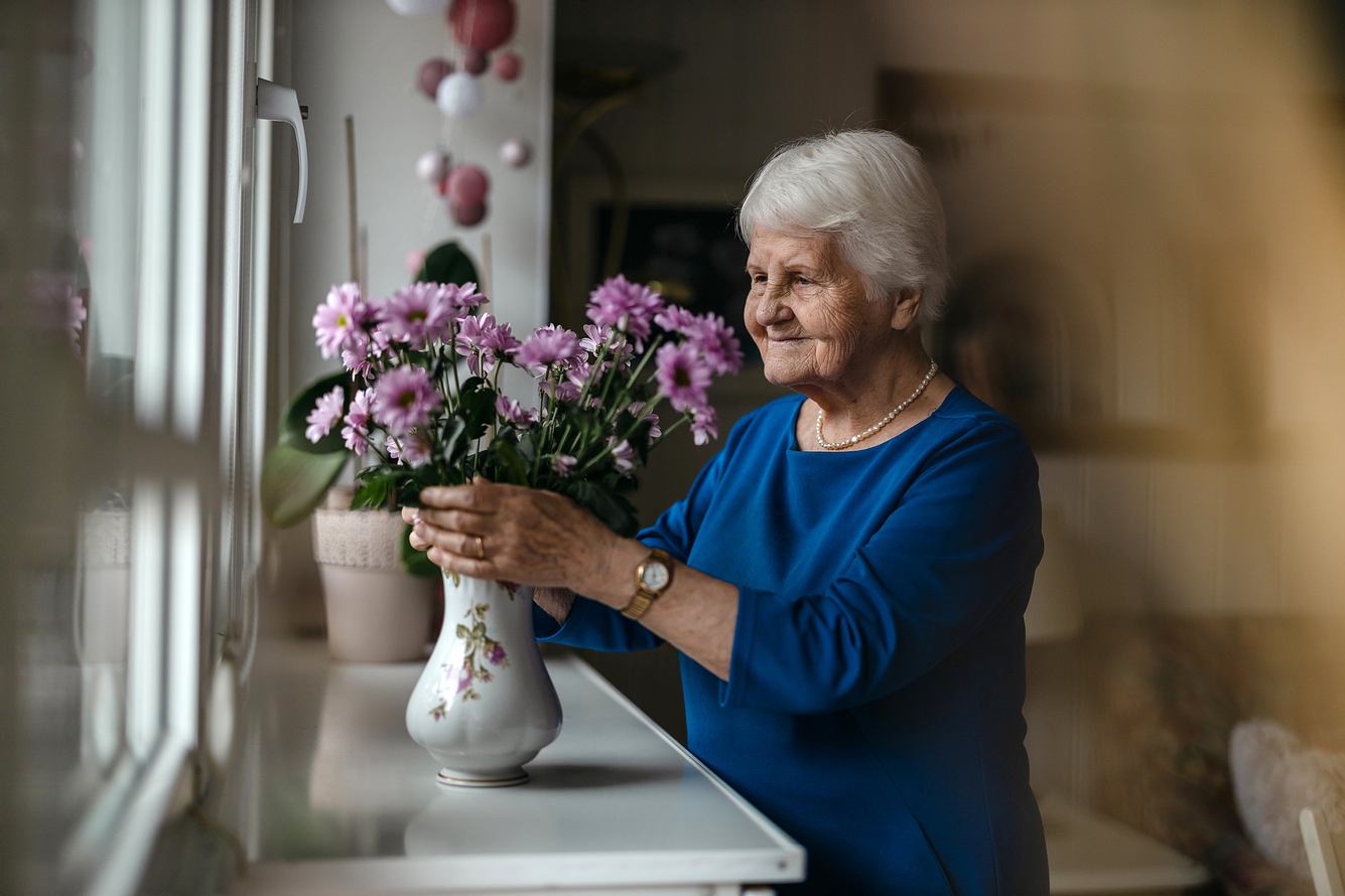 Frau mit Demenz steht am Fenster und berührt Blumenstrauß