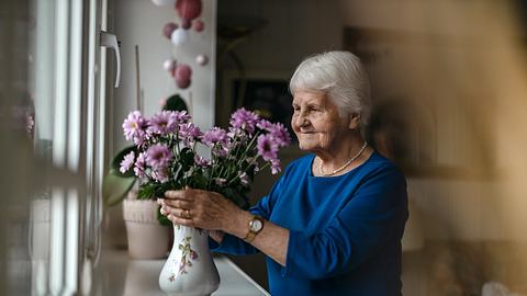 Frau mit Demenz steht am Fenster und berührt Blumenstrauß - Foto: iStock/PIKSEL