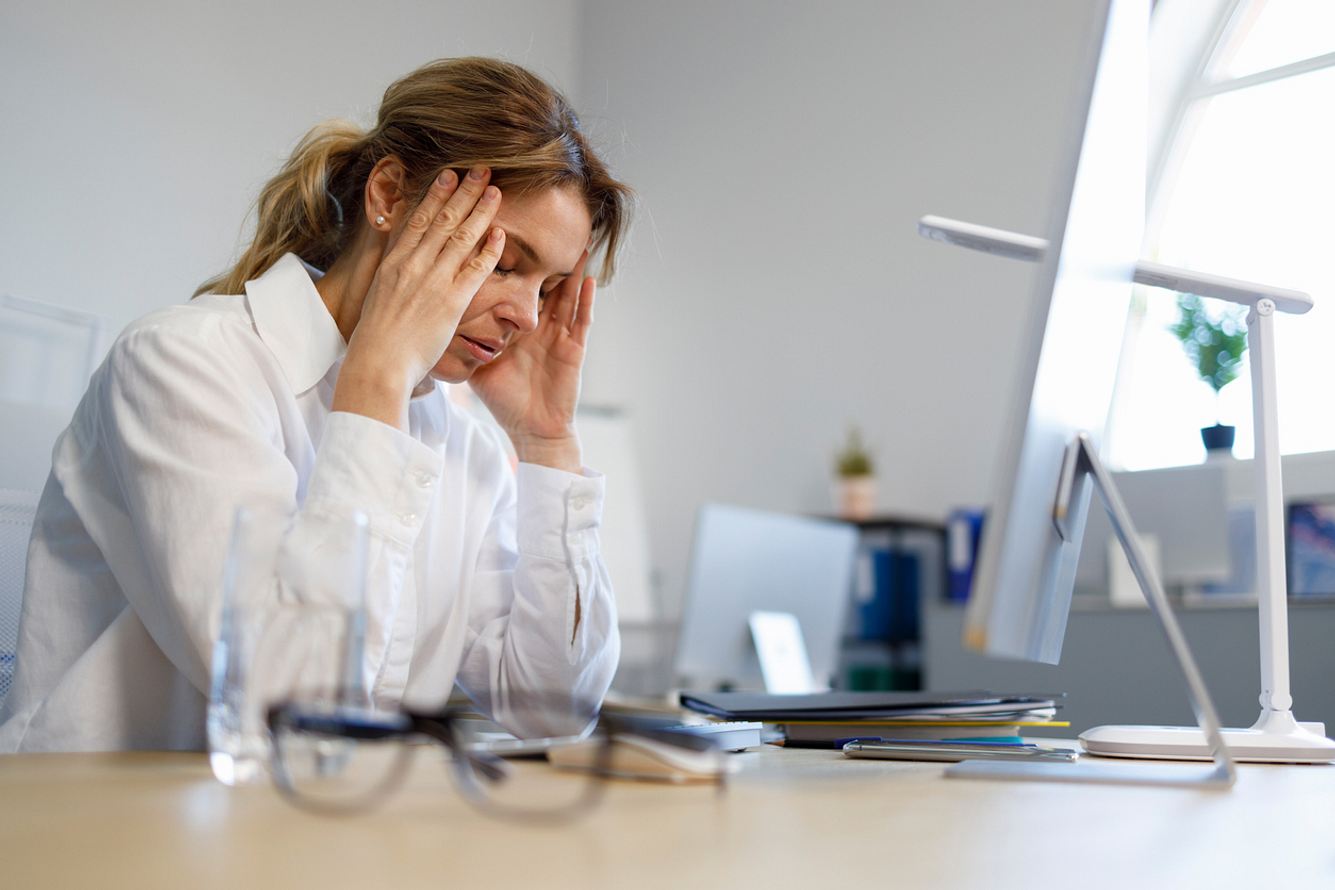 depression-krankschreibung Frau mit weißer Bluse sitzt am Schreibtisch, Augen geschlossen, Kopf in die Hände gestützt