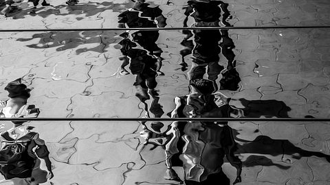 Fußgänger spiegeln sich in einer Wasserpfütze auf einem Gehweg - Foto: Niall_Majury