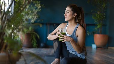 Frau in Sportkleidung sitzt auf einer Yoga-Matte und trinkt einen Smoothie - Foto: iStock/Hispanolistic