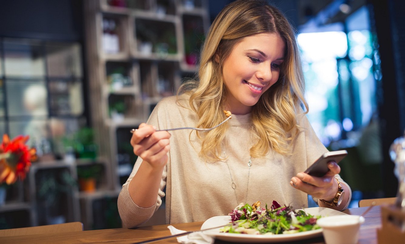 Junge Frau isst Salat und checkt ihr Handy