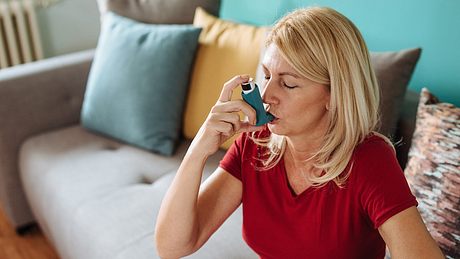 Frau sitzt auf einem Sofa und verwendet ein Asthmaspray - Foto: iStock/drazen
