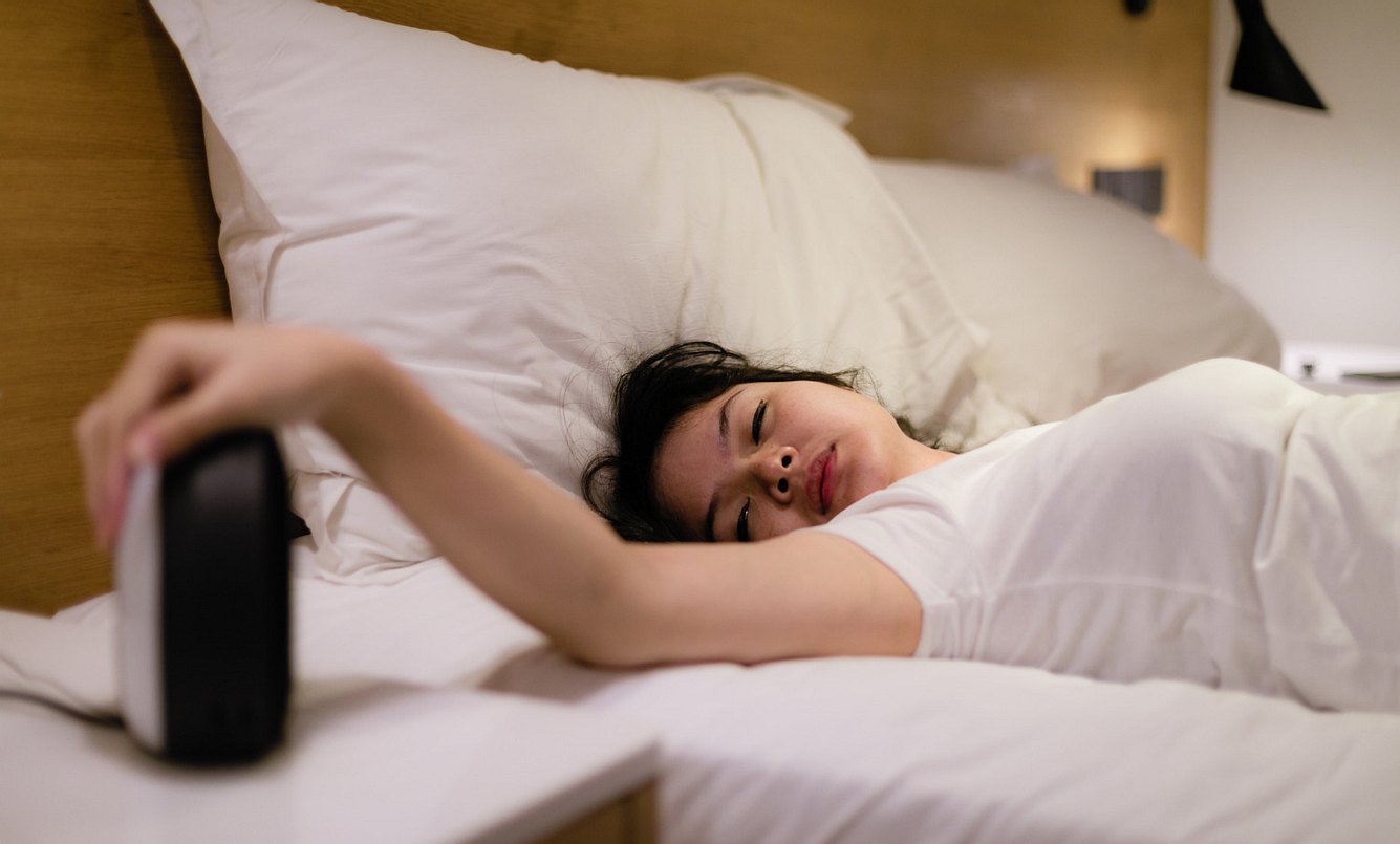 Eine Frau liegt müde im Bett und schaltet ihren Wecker aus