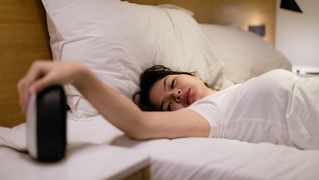 Eine Frau liegt müde im Bett und schaltet ihren Wecker aus - Foto: iStock/rudi_suardi