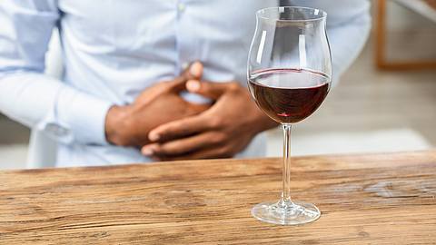 Ein Mann hält sich den Bauch, vor ihm steht ein Glas Rotwein - Foto: IMAGO / Panthermedia