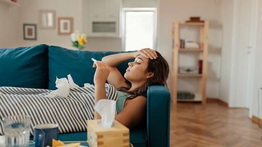 Frau liegt auf der Couch und misst Fieber. - Foto: iStock / PredragImages