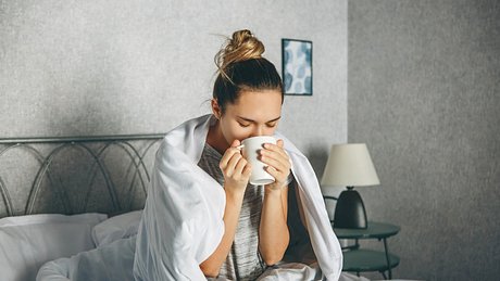 Eine erkältete Frau sitzt auf ihrem Bett und schnäuzt ihre Nase - Foto: iStock_franz12