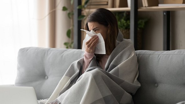 Eine erkältete Frau sitzt auf dem Sofa und putzt sich ihre Nase - Foto: iStock_fizkes