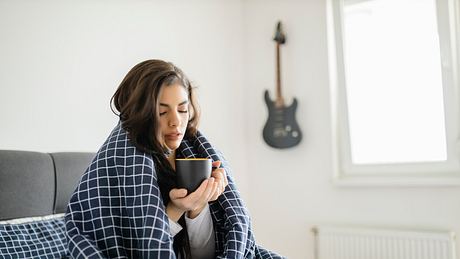 Frau sitz mit Erkältung ohne Schnupfen mit Decke und Tee in Hand und eingekuschelt auf ihrem Bett  - Foto:  iStock/DjordjeDjurdjevic