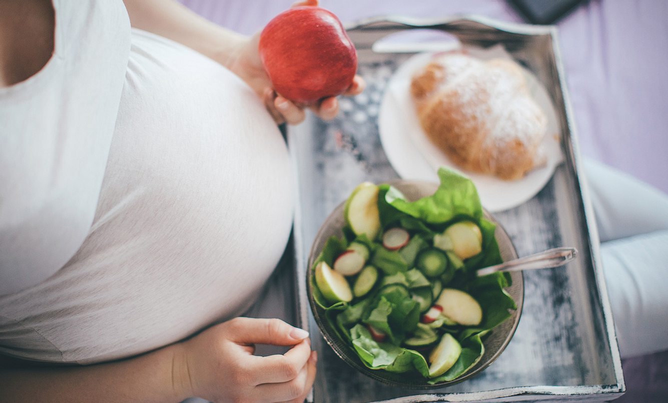 Bauch einer Schwangeren von oben; sie hält einen Apfel in der Hand, vor ihr ein Tablett mit Salat und Croissant