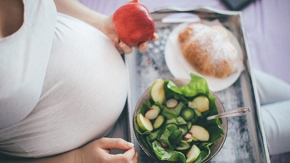Bauch einer Schwangeren von oben; sie hält einen Apfel in der Hand, vor ihr ein Tablett mit Salat und Croissant - Foto: iStock-636752590 Pekic