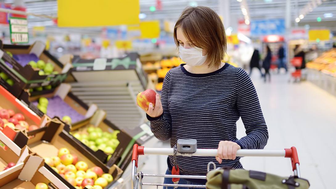 Frau mit Mund-Nasen-Schutz schaut Apfel im Supermarkt an - Foto: iStock/SbytovaMN