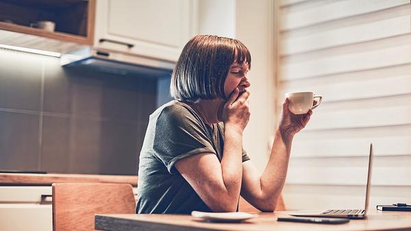 Frau im T-Shirt sitzt mit Kaffeetasse am Tisch und gähnt - Foto: iStock/BitsAndSplits