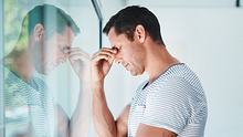 Ein erschöpfter Mann steht vor einem Fenster und hält sich mit geschlossenen Augen und gesenktem Kopf den Nasenrücken.  - Foto: Cecilie_Arcurs / AsiaVision