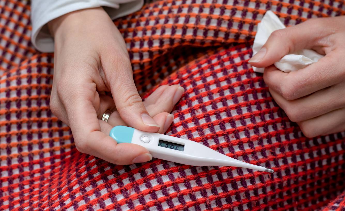 Eine Frau mit einer Decke und einem Fiebertermometer und Taschentuch in der Hand