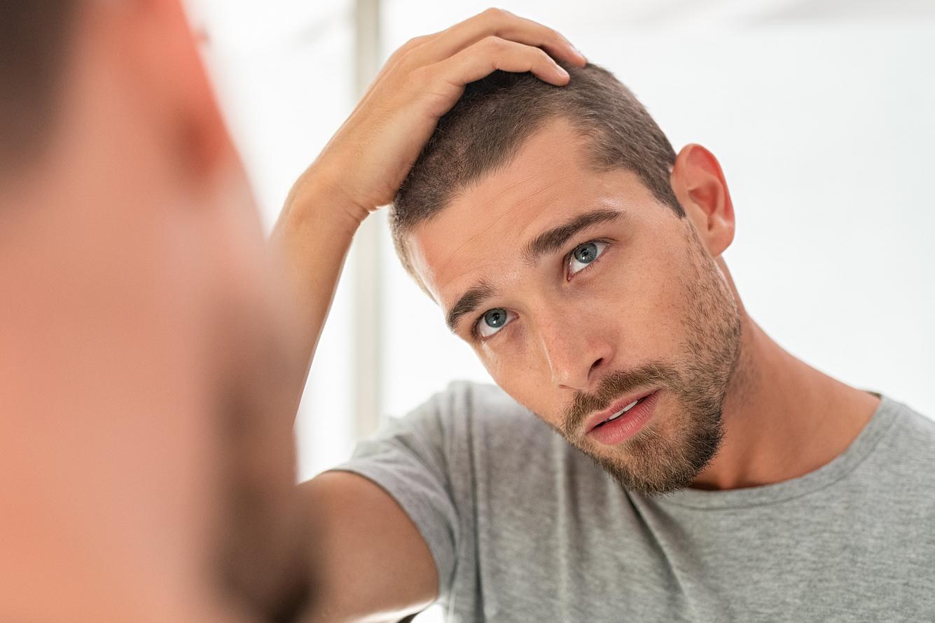 Ein junger Mann sucht im Spiegel nach grauen Haaren an seinem Kopf